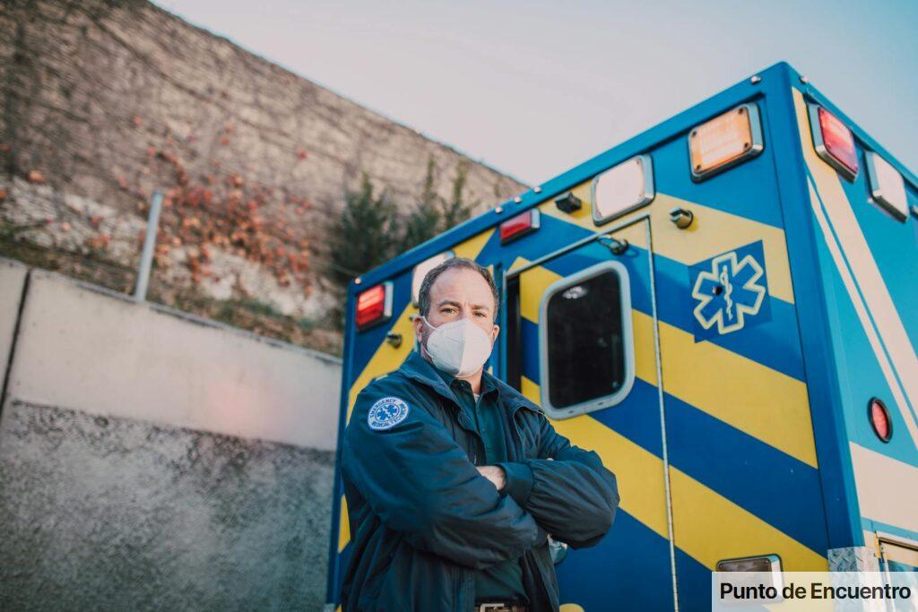 A confident paramedic wearing a mask stands arms crossed by an ambulance.